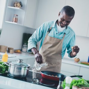 A smiling man in a beige apron cooks in a modern kitchen, pouring oil into a red frying pan with vegetables. Fresh ingredients like lettuce, tomatoes, and onions are on the counter, along with colorful condiments