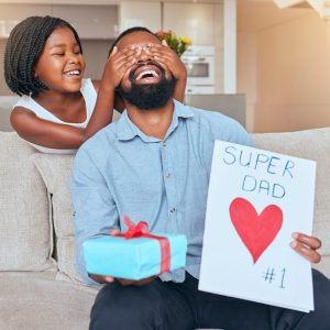 A girl playfully covers her father's eyes while he laughs on a couch, holding a blue gift with a red ribbon and a card that says "SUPER DAD ❤️ #1," perfect for a gift guide for dads.
