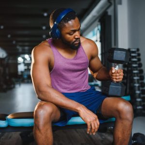 A man in a purple tank top and blue shorts sits on a bench in a gym, intently lifting a dumbbell with his right arm while wearing headphones; a scene showcasing curated gift ideas for health and wellness enthusiast's gift guide.