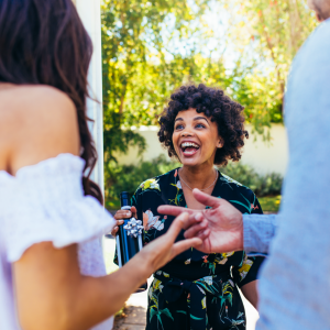 Joyful outdoor gathering with friends, featuring a woman with curly hair and a patterned dress, holding a wine bottle as a gift, surrounded by lush greenery and trees, perfect for a housewarming party gift guide.