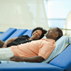 Older couple smiling at each other while lounging on blue deck chairs on a sunny day, enjoying a relaxing treat; a curated gift idea for a leisurely vacation.
