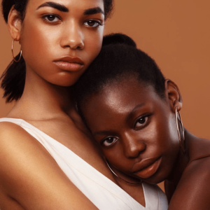 Two women with glowing skin pose together against a warm, neutral background, showcasing the beauty of diverse skin tones. Ideal for a skincare gift guide.